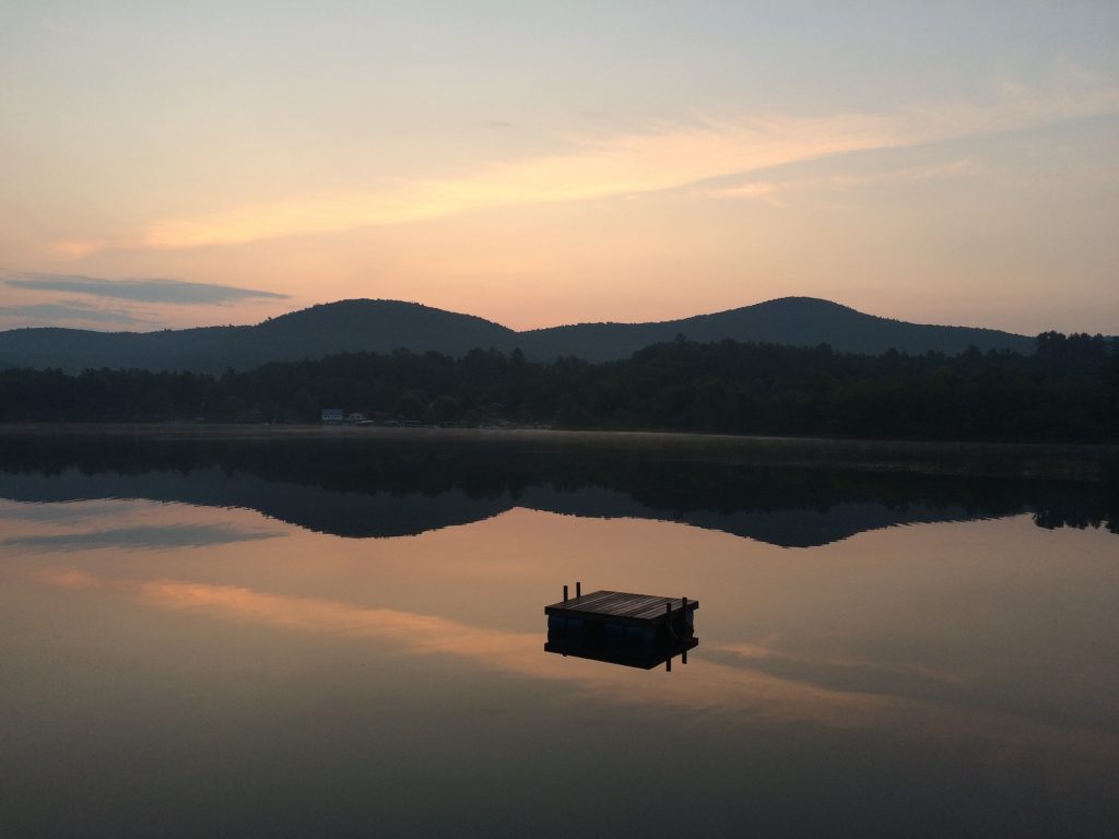 A wooden swim platform in the middle of a lake with the fading color of sunset in the background.