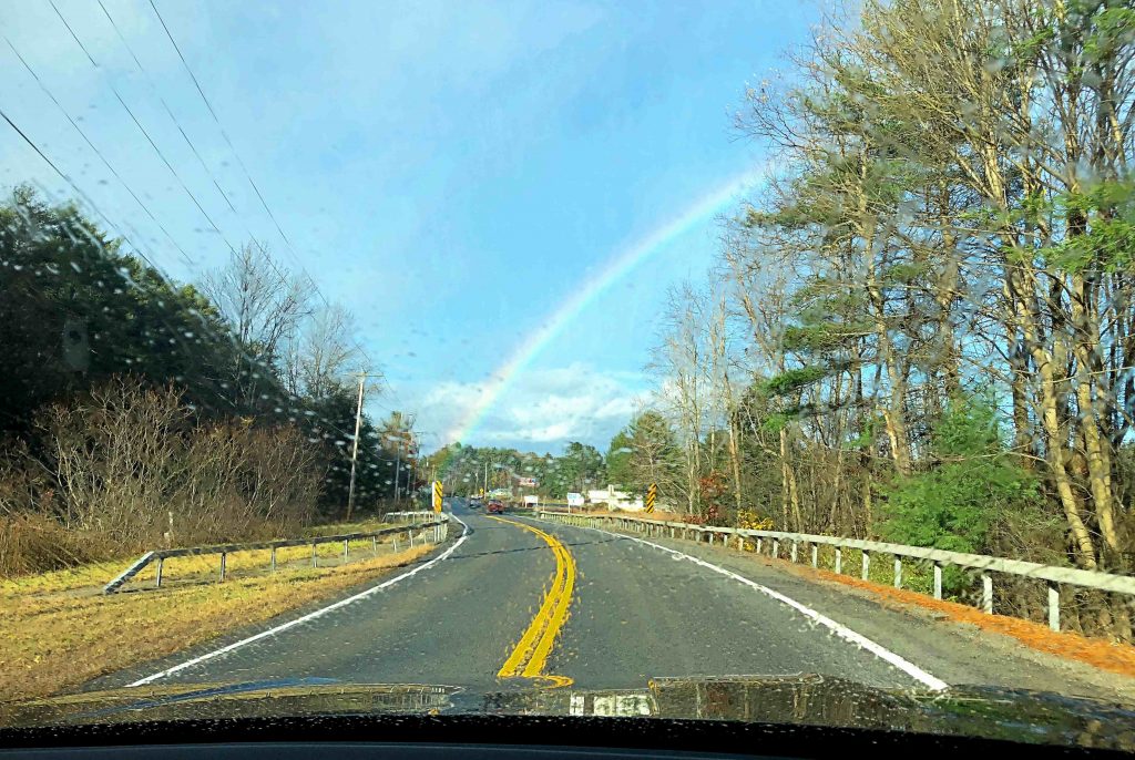 A rainbow across a blue sky seen through the rainy windshield of a black car.