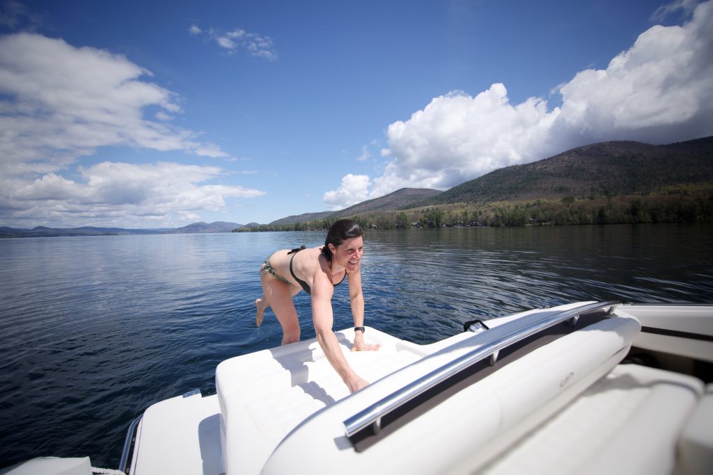 A woman leans over the stern end of a boat to grab a towel. She is in a bikini with a lake behind her.